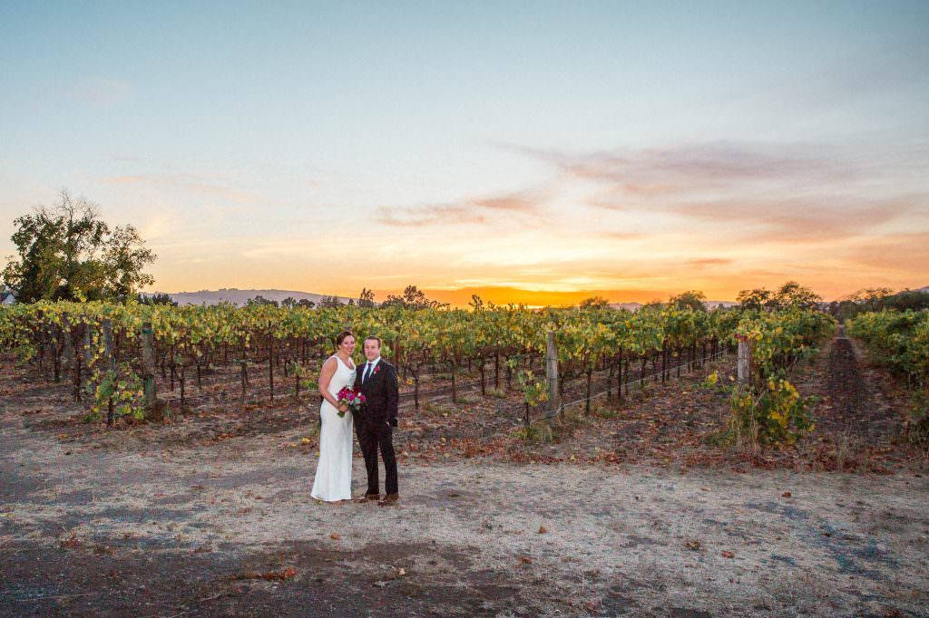 Newlyweds stand in front of a vineyard at sunset